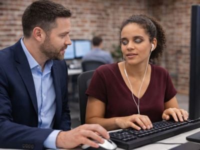 A white man in a navy suit jacket and light blue shirt sitting beside a brown-skinned woman with curly hair pulled back, wearing a maroon top and wired earphones. They are at a desk with a computer keyboard and mouse in front of them, and both are facing slightly toward each other as if discussing something on the computer.